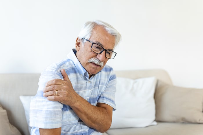 Elderly man sitting on a couch, holding his painful shoulder.